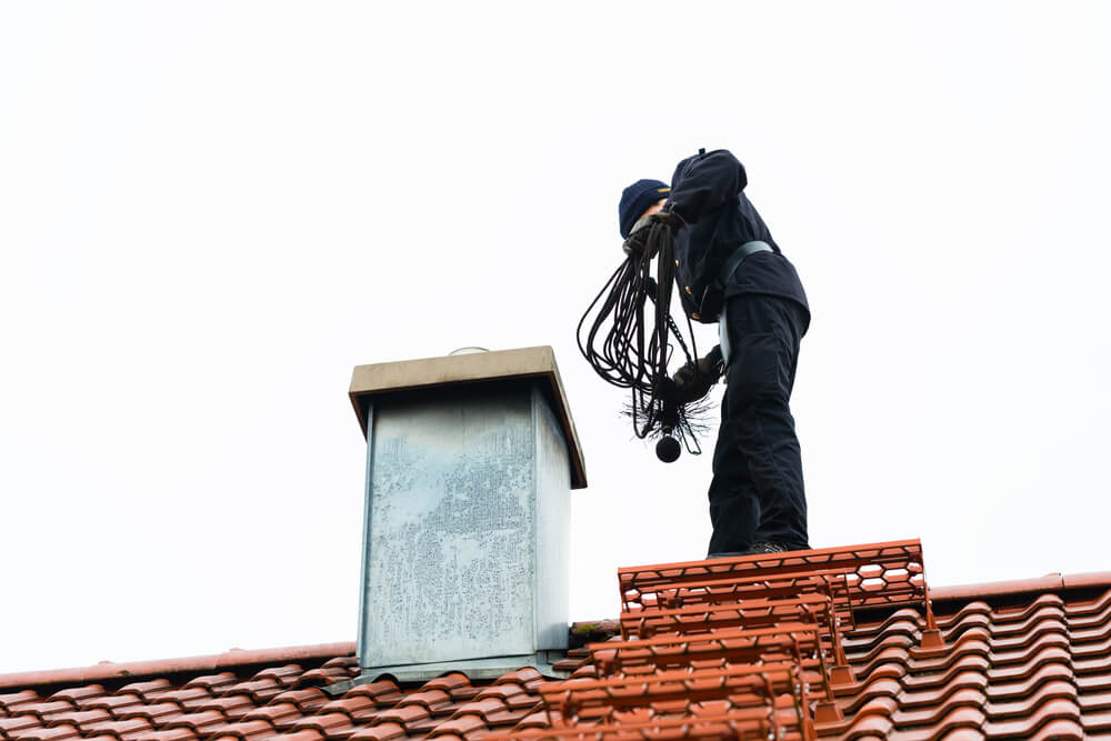 Stock photo of tech with brush looking in the top of a chimney.  The roof is made of tile.