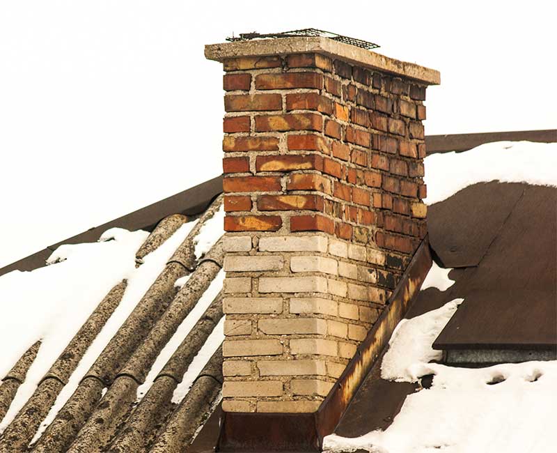 Older chimney with spalling. Chimney has brown brick on top and white on bottom.  The roof is slate and has snow on top.