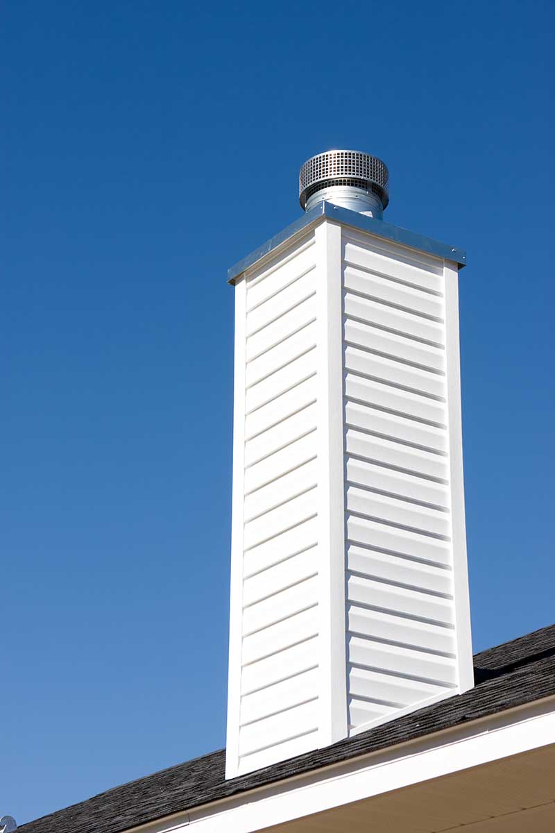 Stock photo of white prefab chimney with stainless steel chase cover sitting on shingle roof with beautiful blue sky in the background.