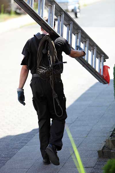 Tech walking down sidewalk carrying ladder with orange flag on it.  He also has his safety equipment on.