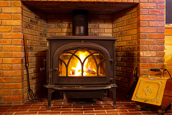 Stock photo of wood burning stove sitting inside a large fireplace.  brass kindling holder to the right and poker to the left.  The surround and hearth is all red brick.